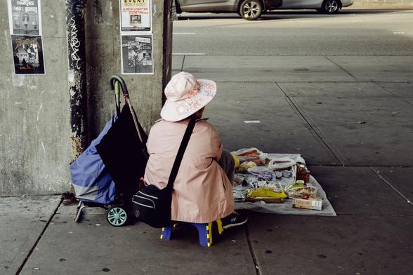 A woman quietly sells groceries on the pavement beneath an overpass.