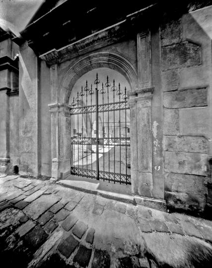 The photograph depicts the courtyard of the Armenian Church in Lviv, captured with a pinhole camera. The image centers on an ornate iron gate set within a stone archway, framed by weathered architectural details. The cobblestone path in the foreground leads the viewer’s eye toward the sunlit courtyard beyond the gate, where soft light and shadow create a tranquil, almost timeless atmosphere. The characteristic softness and slight distortion of the pinhole technique add a dreamlike quality to the historic setting.