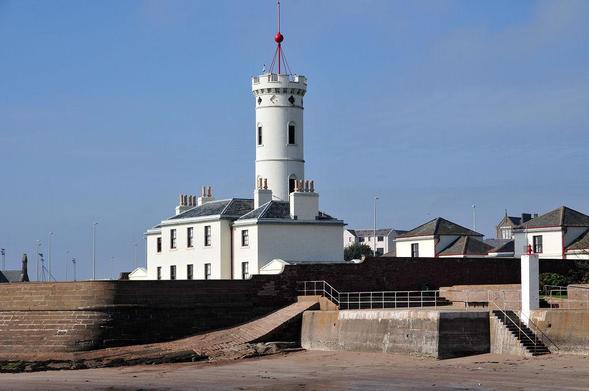 Arbroath's Signal Tower Museum. The image shows a view across a sandy beach at low tide to stone steps and a sea wall built of red stone facing to the left. Beyond it, in the centre of the frame, is a white building with a grey slate roof and lots of chimneys. Rising out of it is a white circular tower looking like a slender rook in a chess game. A pole on its top carries a signal ball. There are the roofs of more buildings to the right. The sky is blue.