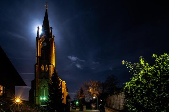 Foto einer grossen Kirche in der Nacht. Man sieht eine Gasse sie zum Gebäude führt, rechts Bäume und ein Zaun, links sind Teile eines Hauses zu erkennen.
Hinter einem der Kirchtürme steht der Mond, der die ganze Szene beleuchtet. Der Himmel ist fast wolkenlos, ein paar Sterne sind auch zu erkennen.