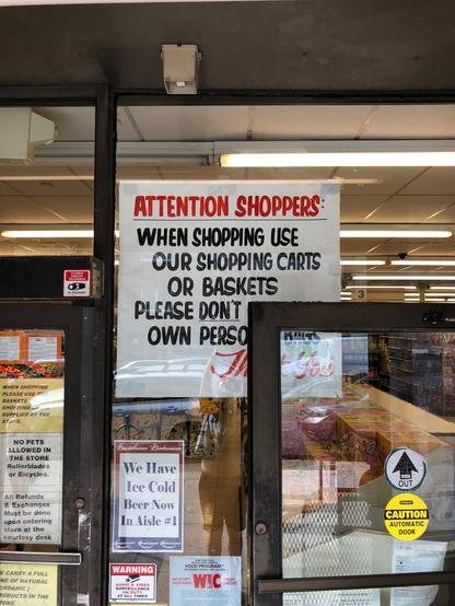Hand-painted sign reading "Attention Shoppers: When shopping use our shopping carts or baskets. Please DON'T [use your] own perso[nal] bags. Th[an]k You"