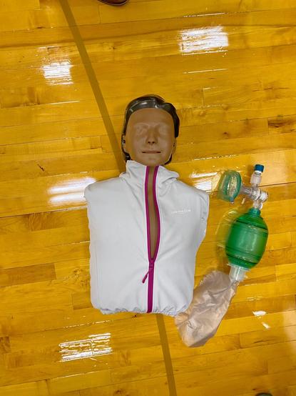 Color image of a training mannequin torso on a gym floor, sometimes nicknamed Ambu Man or Resusci Manny. Image shows the armless torso male mannequin wearing a white training jacket with a purple zipper, tan/brown skin tone, black hair, lying on a wood strip gymnasium floor, with an ambu bag medical device lying next to him, green inflation pump and face mask, clear tubing and inflation bag.