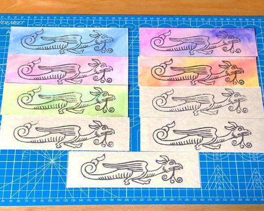 several block printed bookmarks laid out on a blue cutting mat.
five on watercolor backgrounds, four on parchment patterned card stock.

the block print is of a long wyvern like creature with feathery wings a corkscrew tail, and a beak like mouth with curls hanging out of it like it's eating something.