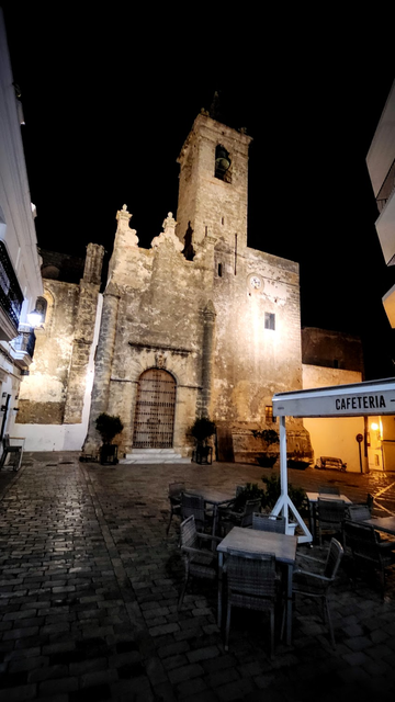 Ein Platz vor einer Kirche in einem alten Ortsteil. In der Nacht ragt die Kirche in den dunklen Himmel. Das Kopfsteinpflaster liegt verlassen da. Im Vordergrund sind die Tische einer Bar und Cafeteria leer und geputzt für den nächsten Tag.