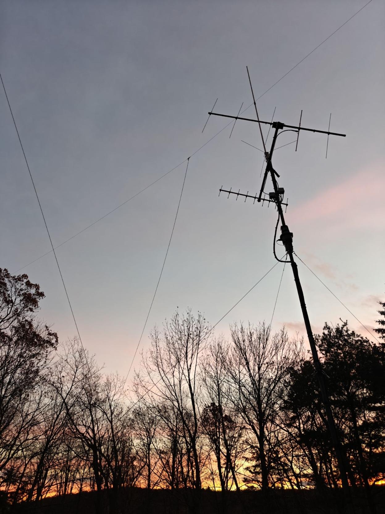 Yagi antennas for satellites, a vertical for VHF/UHF, and a HF doublet against a slightly cloudy sky with a backdrop of trees through which the ligt of the setting sun is visible.