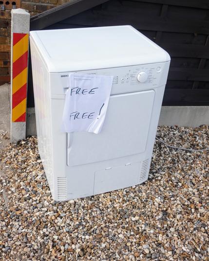 A white clothes dryer is on gravel near a building's stairs. It has a white piece of paper on it saying:
"FREE
FREE!"