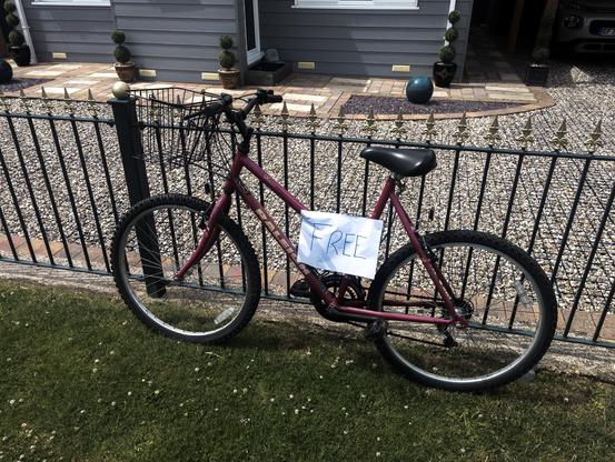 A Raleigh bike has a piece of paper attached to it saying "FREE". It is leaning against a fence, in a greassy area in front of a graveled front drive of a house.