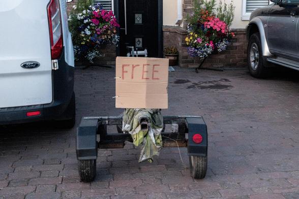 A trailer in the front drive of a house has a piece of cardboard attached to it with cableties saying "FrEE"