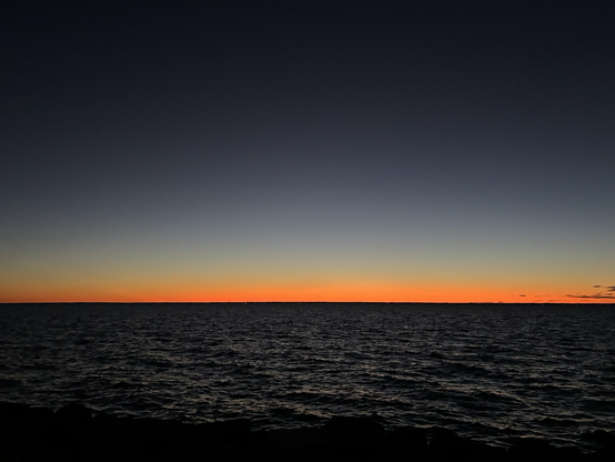 twilight over the water — the sun has just slipped below the horizon, leaving behind a brilliant band of orange and gold that fades smoothly into a deep indigo sky.