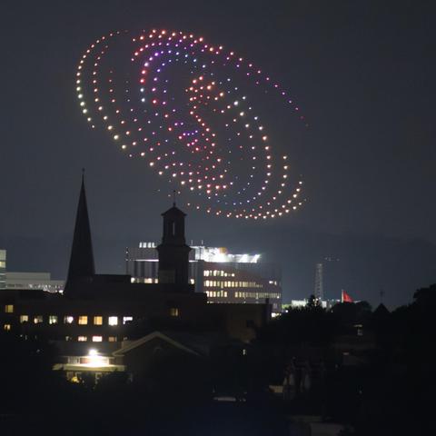 A nighttime scene featuring a spiral pattern of colorful lights in the sky, likely from a drone display, over a city skyline. Silhouettes of buildings and church spires are visible in the foreground, contrasting against the illuminated sky.
