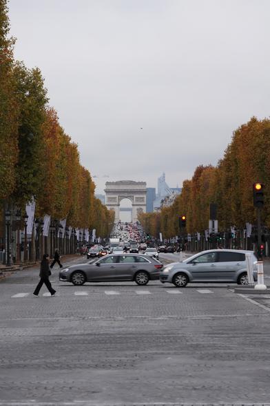 Avenue des Champs-Élysées through a long lens, which compresses the nearly 2km and shows all the traffic coming. Many people have taken this picture. Meanwhile two pedestrians are crossing the road, as well as two cars. The Arc de Triomphe at the end of the road is seen between the rows of trees at the side of the road.