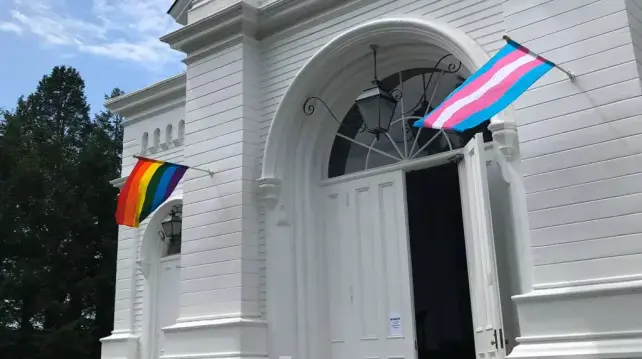 church hosting spanish classes. pic shows fancy churcj door, open, with pride and trans flags on either side