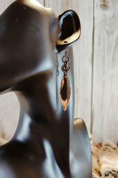 Boucle d'oreille composée majoritairement d'une grande écaille en acier inoxydable, et dont le bord est dentelé. On ne voit qu'une boucle de la paire, portée sur un support.