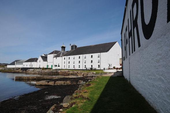 Laphroaig Distillery. The image shows a view along the front of a white warehouse which has, in the top right of the frame the letters LAPHRO and part of the A visible. The shore is to the left of it and a little around the shore is a range of white buildings of up to three storeys in height, topped off by a distillery pagoda. The sky is blue.