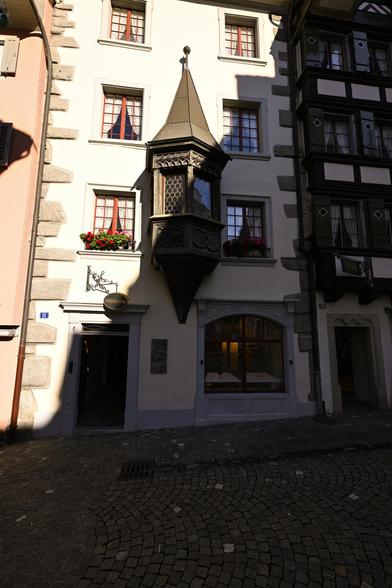 The image shows a narrow street view of two buildings with cobblestone pavement. On the left is a pale, multi-story building with several windows, each adorned with red flower boxes. Protruding from the building is an ornate, dark-colored, multi-tiered structure, resembling a small tower with openings. To the right is a darker building with a white facade and black detailing; the windows are rectangular and arranged in neat rows. The image appears to be taken during daylight, with shadows cast on the pavement. Number “31” is visible on the left building and “33” on the right building.

Provided by @altbot, generated privately and locally using Gemma3:27b

🌱 Energy used: 0.110 Wh