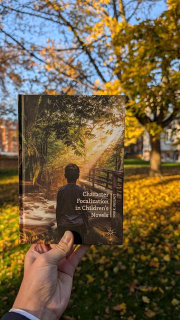 Holding the book "Character Focalization in Children's Novels" in front of a yellow-leafed tree