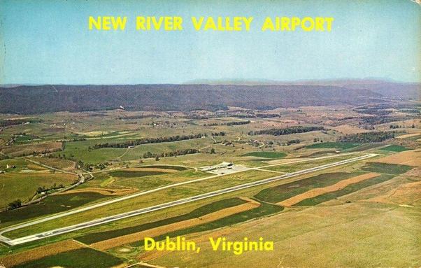 This vintage postcard shows an aerial view of New River Valley Airport in Dublin, Virginia. The photograph captures the airport and its surrounding countryside bathed in soft daylight. A single long runway stretches diagonally across the lower half of the image, bordered by neatly divided patches of farmland in shades of green, gold, and brown. Near the center sits the small airport terminal and parking area, with a few structures and paved roads visible. Beyond the airfield, the land rolls gently into the distance, giving way to tree-covered hills and a deep blue mountain range under a clear, bright sky. Large yellow text across the top reads “NEW RIVER VALLEY AIRPORT,” and at the bottom, “Dublin, Virginia.” The image evokes a sense of open space, rural tranquility, and mid-century optimism in regional aviation.