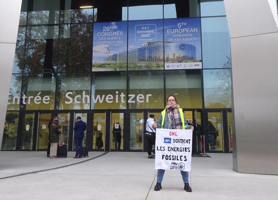 Une activiste en gilet jaune tient une banderole qui dit "GNL : Axa soutient les énergies fossiles" devant l'entrée du palais des congrès à Strasbourg. Derrière elle, des participant·es s'apprêtent à entrer dans le bâtiment. Au-dessus de l'entrée, on voit un grand affichage annonçant ce 26e congrès européen des agent·es AXA.