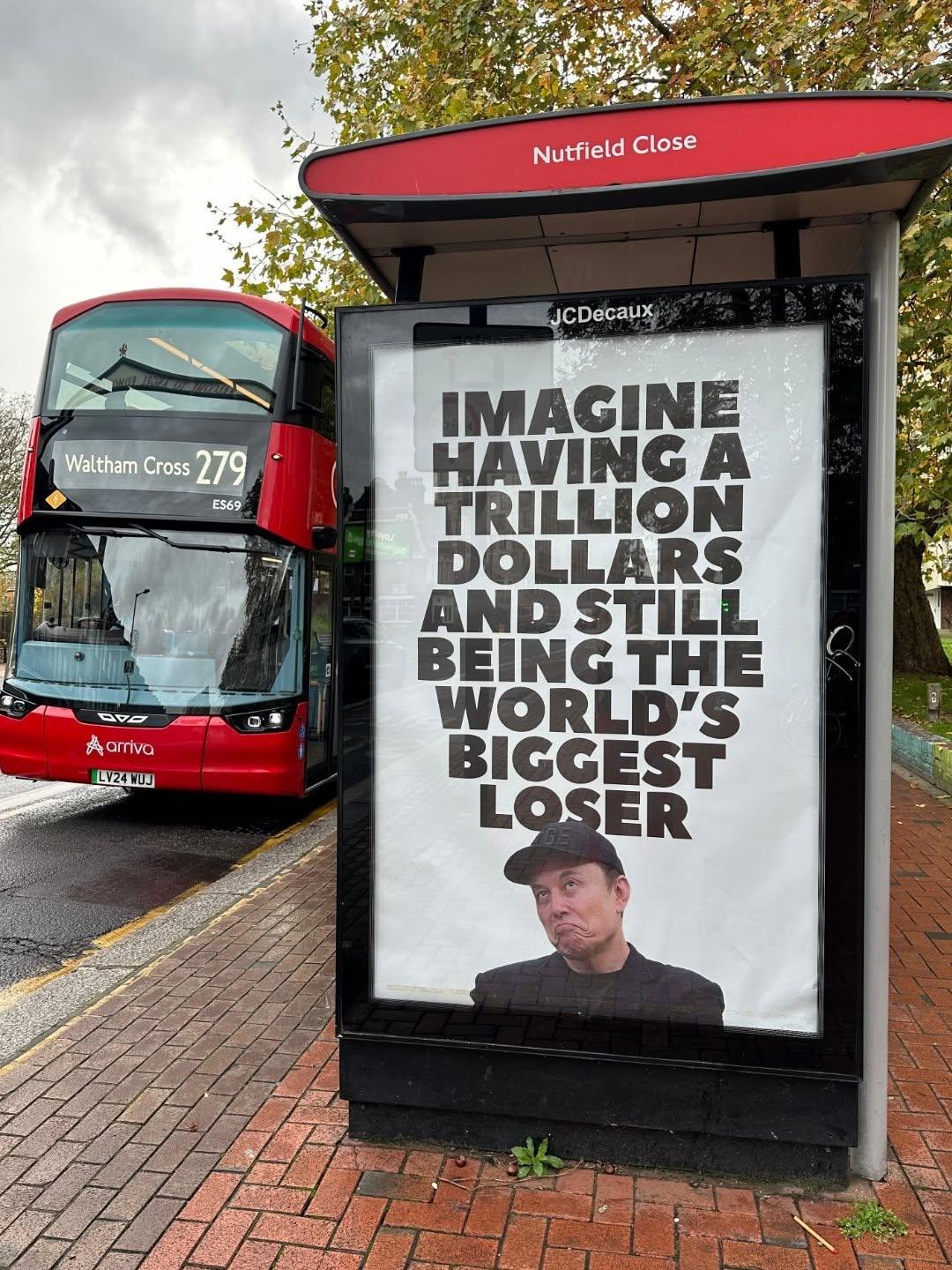 A satirical poster at a London bus stop on Nutfield Close features a bold message reading, “IMAGINE HAVING A TRILLION DOLLARS AND STILL BEING THE WORLD’S BIGGEST LOSER,” above an image of a man making a sarcastic expression. A red double-decker bus with the destination “Waltham Cross 279” passes by on the street, and autumn trees line the background. Created by the anonymous artist collective everyonehateselon_, this piece humorously critiques billionaire culture and public perception of extreme wealth.