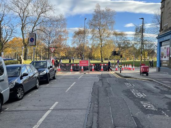 Urban street ended in a T junction, closed to cars by cones and signs. A park with autumnal trees in the backdrop. The sky is blue with light clouds