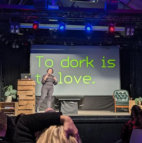 Astrid stands on stage in front of a large screen displaying the phrase “To dork is to love.” in bright green text. The venue is dimly lit with blue and red lights, and the seated audience listens attentively.