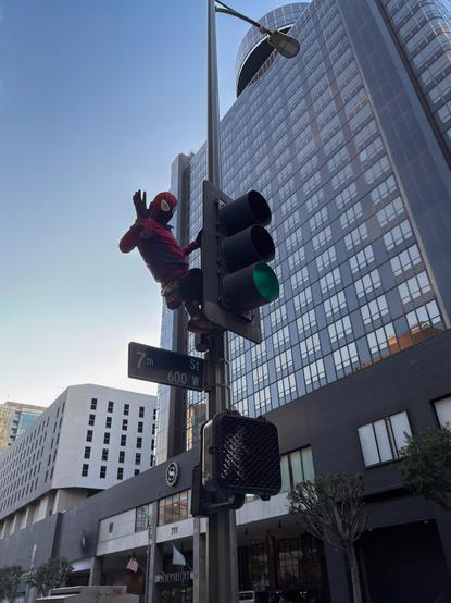 A person in a full Spider-Man costume, who has climbed up a traffic light pole and is waving at the camera from above.