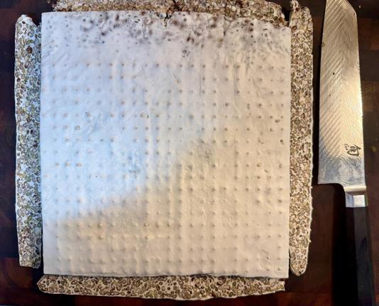 A square white slab of tempeh with a matrix of small dots from the perforations in the plastic bag that held the batch. The four rounded edges have been cut off and show the mix of lentils and barley embedded in the white matrix of the tempeh mycelium. At the side is the knife that was used.