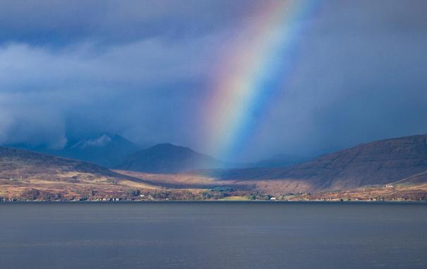 A rainbow in front of very grey mountains with some grey sea in the foreground.