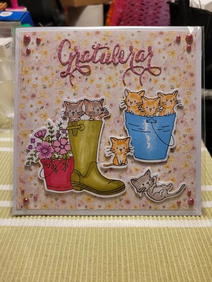 Grattiskort med katter i ebän grön stövel brevid en rödrosa hink med rosa blommor.
2 kattungar en röd och en grå sen en blå hink med katter 
Blommig bakgrund i rosa och gult 
Gratulerar utstansat i blommigt rosa och vitt papper.