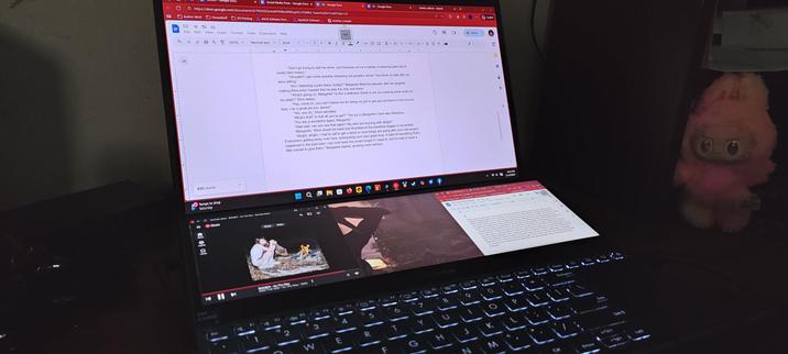 An open laptop with a writing project on the screen, Eve from the game Stellar Blade on the secondary display, and a Loyalty Labubu watching the keyboard.