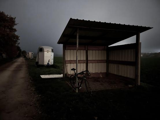 Eine dunkle Landschaft mit einem Feldweg, an dem zwei Anhänger geparkt sind. Im Vordergrund ist ein offener Unterstand zu sehen, an den ein Fahrrad gelehnt ist, während ein weit entferntes Feld und die schlechte Sicht aufgrund von Nebel eine stimmungsvolle Atmosphäre schaffen.