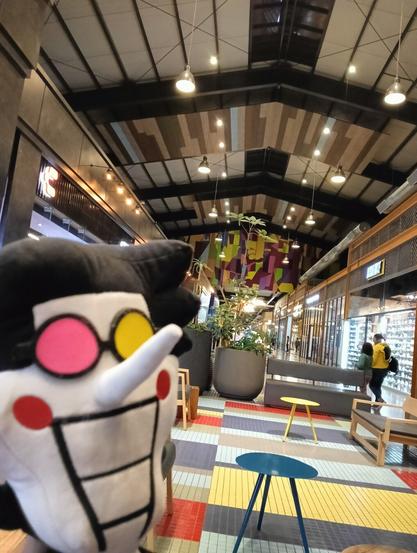 The image shows the interior of a shopping mall or similar commercial space, with a plush toy of a character with yellow and pink eyes, black hair, and red circles on its cheeks in the foreground. The background features a walkway with shops on either side, benches, plants in large pots, and colorful tiled flooring. The ceiling has a modern design with exposed beams and hanging lights.