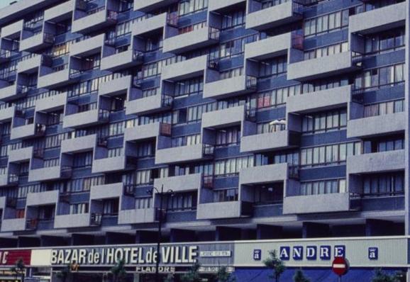 Photographie en 1970 d'un immeuble moderne avec des balcons et fenêtres en zigzag. Au rez-de-chaussée les enseignes de commerce Bazar de l'hôtel de ville et André