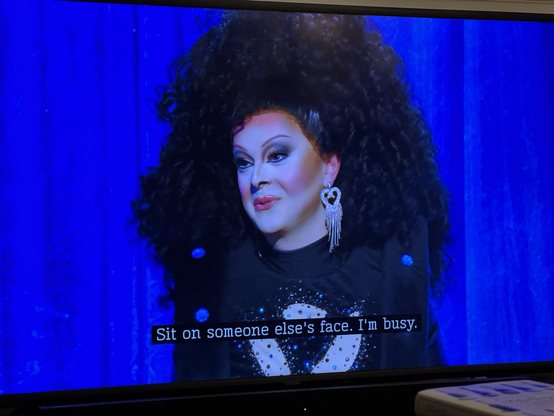 On the set of Snatch Me Out, Bonnie Ann Clyde, appearing as Chair (long story) in full curly black hair and dress say “Sit on someone else’s face, I’m busy