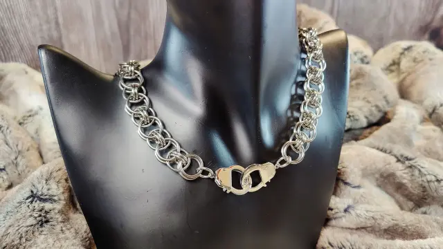 Collier en maille utilisant des anneaux un peu plus grand que la moyenne, et le fermoir est une paire de petites menottes (fonctionnelles, mais sans clés). Photographié ici sur un présentoir.