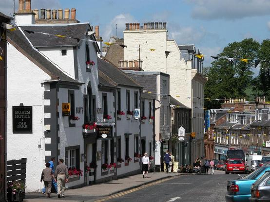 The High Street in Melrose. The image shows along a street crossed by lines of bunting that descends a hill. The buildings on the left occupy most of the frame and comprise mainly white two or three-storey structures. The nearest is signed Burts Hotel. The buildings on the right are visible as the road curves left at the bottom of the hill and are mainly of stone. The scene is in sunlight.