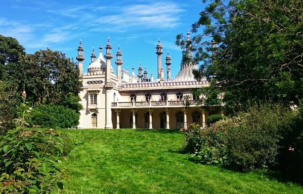 A colour photograph of the Royal Pavilion in Brighton, an ornate 19th-century palace featuring distinctive Indo-Saracenic architectural style. The building is characterised by its intricate domes, minarets, and elaborate detailing. The structure is set against a backdrop of lush greenery, with a well-manicured lawn in the foreground and trees framing the scene. The sky is clear and blue.