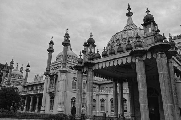 A black-and-white photograph of the same Royal Pavilion in Brighton, focusing on its grand façade. The image highlights the architectural details, including the domes, minarets, and arched windows. The perspective is closer, allowing for a detailed view of the ornate carvings and structural elements. Two individuals are visible near the entrance, providing a sense of scale to the imposing building. The sky is overcast.