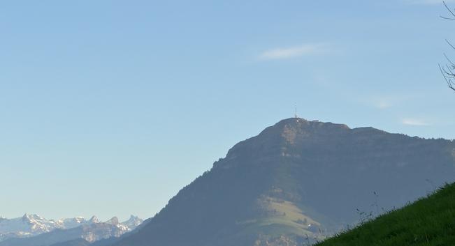 Blick von einem Hügel auf der gegenüber liegenden Talseite auf einem Berg (die Rigi) mit Sendemast. Links vom Berg sind Schneebedeckte Berge zu sehen im Hintergrund