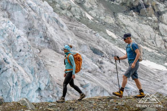 Two hikers wearing blue shirts and backpacks walking along a rocky trail in front of a massive glacier, showcasing the scale and beauty of the alpine landscape.
