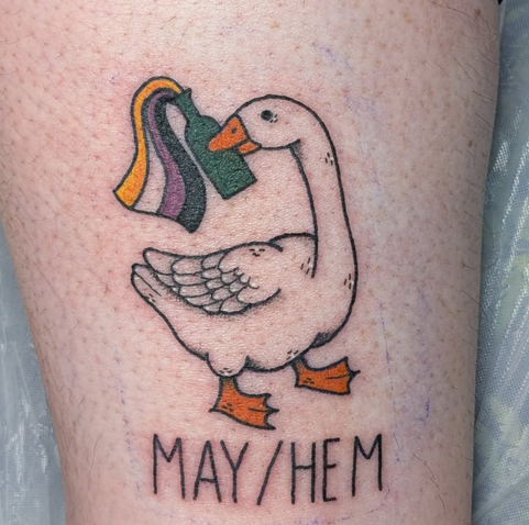 Photo of a coloured tattoo picturing a goose holding a molotov cocktail in their beak, of which the wick is nonbinary flag coloured. Below it, the text "MAY/HEM" is tattooed.
