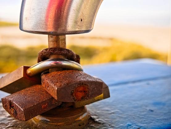 Several heavily rusted padlocks stacked on top of each other, attached to a silver handle. In the background (out of focus): dunes, sand, sea.