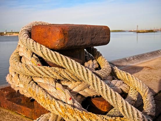 A bright rope is wrapped several times around a rusty bollard. Behind it is a harbour.