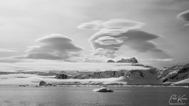 Photographie monochrome en noir et blanc, présentant de nombreux nuages lenticulaires au-dessus d'une baie partiellement encombrée de glace et bordée par des montagnes, entièrement enneigées et englacées.
Il ne fait pas très chaud, même si c'est le printemps en Antarctique.