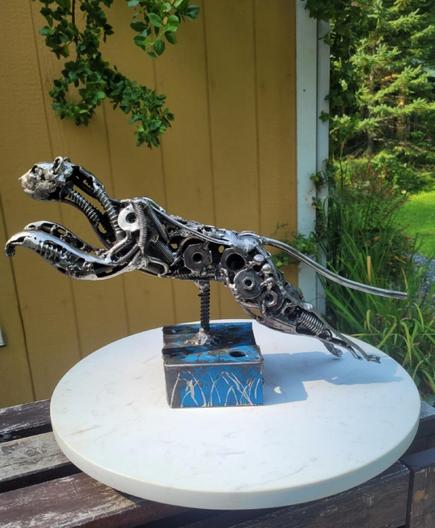 Pressé 
Un petit félin fait de rebut d'acier saute par dessus une structure d'acier de forme rectangulaire. Il est composé de boulons, washers, clous, vis, mailles de chaînes et j'en passe. Il est dehors au soleil sur un socle à gâteau blanc.

100% rebut