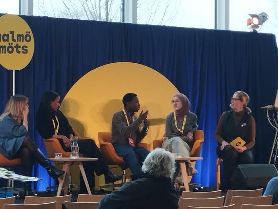 A group of four people are seated on a stage in front of a blue backdrop featuring a large yellow circle. The individuals are positioned on orange armchairs arranged in a semi-circle. One person, positioned to the left, is facing away from the camera and wearing a dark jacket, while another person to the right is wearing a head covering. The person in the center is actively gesturing with their hands while speaking. In the foreground, rows of empty seats are visible, suggesting an audience setting. The text "Globalmots" is visible on the upper left corner of the image.

Provided by @altbot, generated privately and locally using Gemma3:27b

🌱 Energy used: 0.099 Wh
