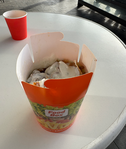 Das Bild zeigt eine geöffnete Dönerbox mit Pommes und Tzatziki-Sauce, die auf einem weißen Tisch steht. Daneben befindet sich ein roter Becher.