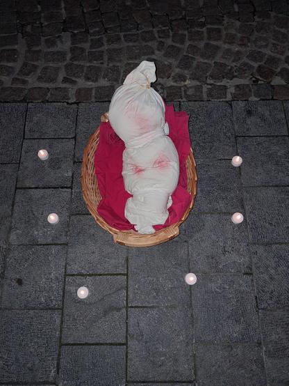 Foto van een babypop in met rode verf besmeurde lijkwade. De pop ligt in een mandje op een rood kussen, erom heen staan 6 theelichtjes.