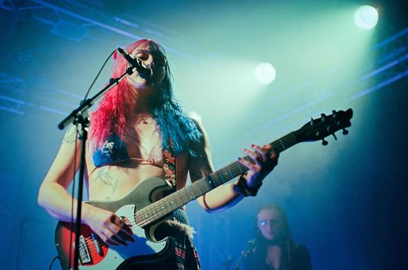 Jasmine4t on stage singing and playing her electric guitar. She has pink and blue hair. She is wearing a bikini and a skirt.
