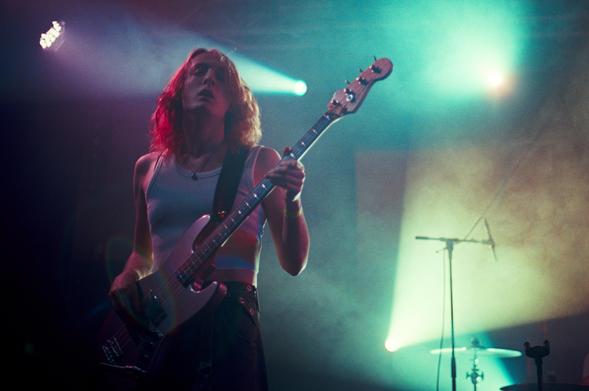 The bassist of Jasmine.4.t playing on stage. They are wearing a white tank top and a black cargo pants.